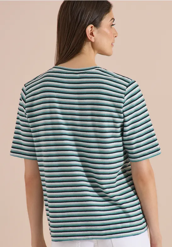 Femme de dos portant un t-shirt rayé blanc et turquoise sur fond beige.