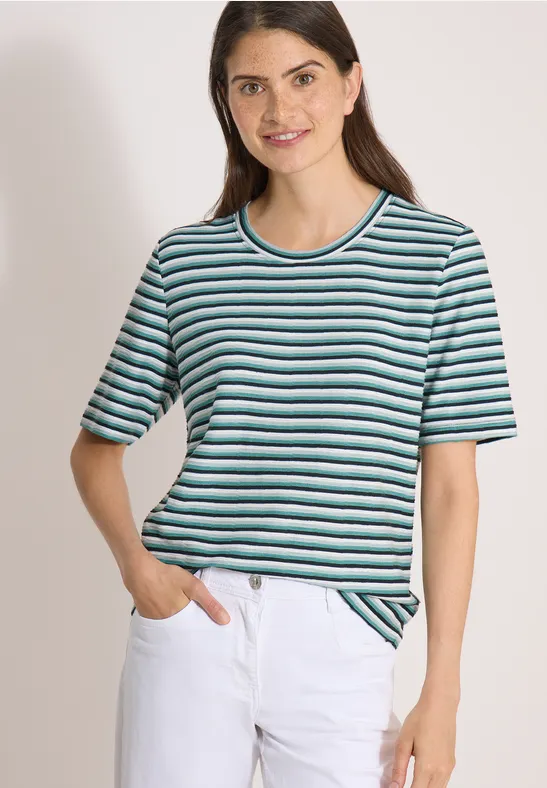 Une femme porte un t-shirt à rayures bleues, vertes et blanches à manches courtes et un jean blanc.