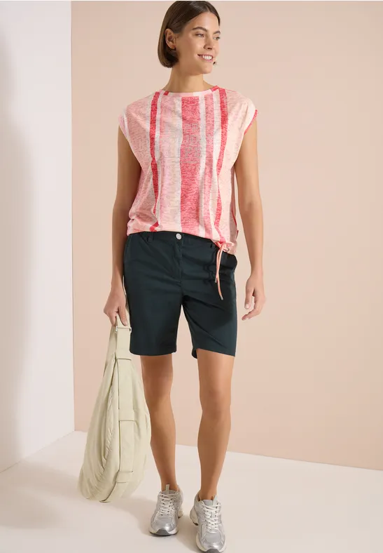 Frau mit rosa-weiß gestreiftem Top, schwarzen Bermudashorts, grauen Sneakers und grünem Beutel.