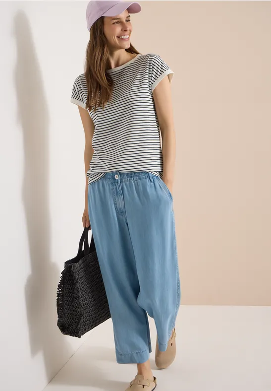 Frau in gestreiftem T-Shirt, hellblauen, weiten Hosen, rosa Cap und Korbtasche lehnt an Wand.