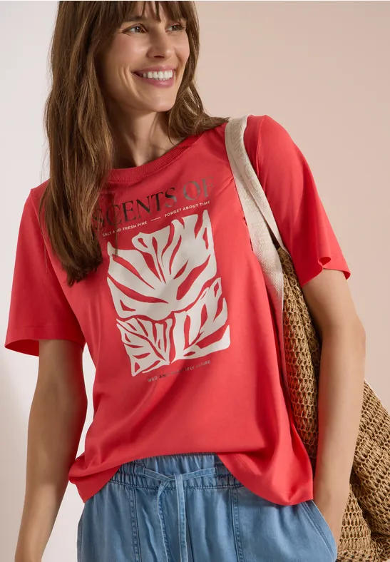 Femme portant un t-shirt rouge à motif de feuilles blanches, un jean clair et un sac en osier sur l'épaule.