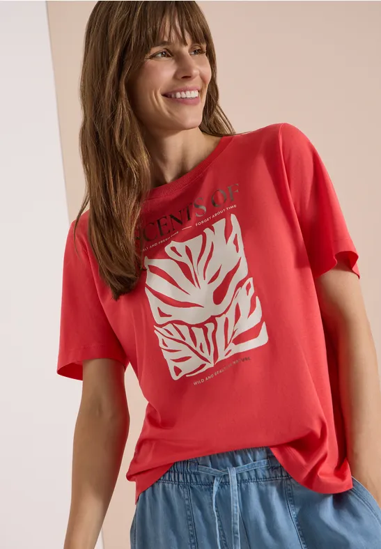 Femme en t-shirt rouge à motif blanc stylisé et jean bleu, posant devant un arrière-plan neutre.