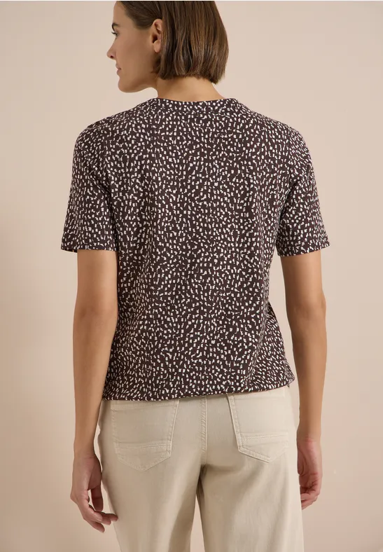 Femme de dos portant un t-shirt noir à petits motifs blancs et un pantalon beige clair sur fond neutre.