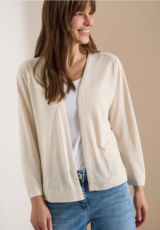 Frau trägt beigefarbene Strickjacke, weißes T-Shirt und blaue Jeans vor schlichtem Hintergrund.