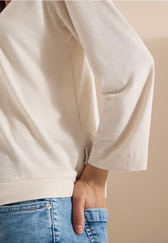 Person trägt cremefarbenes Oberteil und blaue Jeans, Hand in der Gesäßtasche.