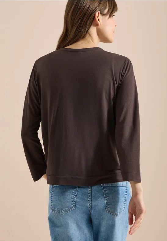 Frau von hinten in schwarzem Langarmshirt und blauer Jeans vor hellem Hintergrund.