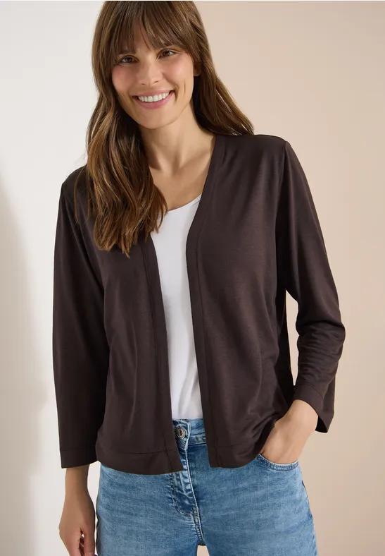 Frau in schlichter dunkler Strickjacke über weißem T-Shirt und blauer Jeans, Hand in Hosentasche.