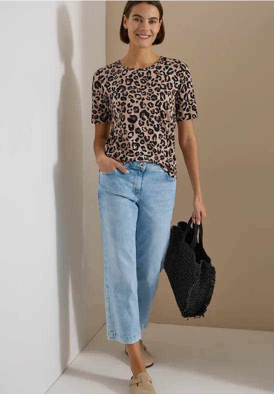 Femme en t-shirt léopard, jean bleu clair et sac tressé noir sur fond neutre.