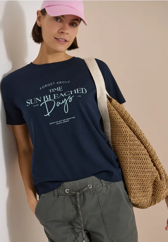 Person mit dunkelblauem T-Shirt „Sun Bleached Days“, grauen Cargohosen und Strohtasche.