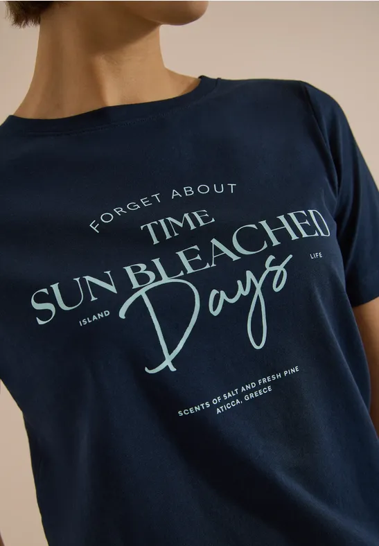 Eine Person trägt ein dunkelblaues T-Shirt mit dem Schriftzug ‚Forget About Time Sun Bleached Days‘.