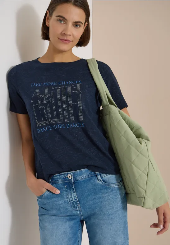 Frau in Jeans, blauem T-Shirt mit Schriftzug und grüner, gesteppter Umhängetasche.