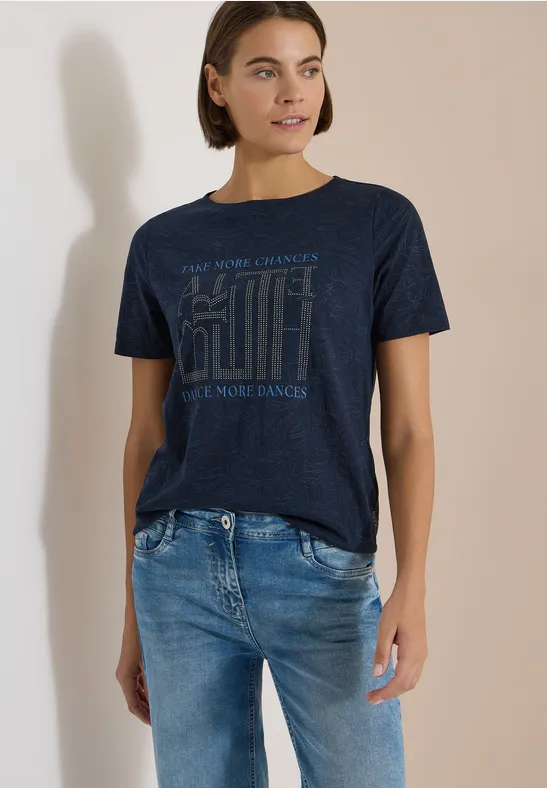 Frau in neutraler Umgebung trägt dunkles T-Shirt mit Print und blaue Jeans.