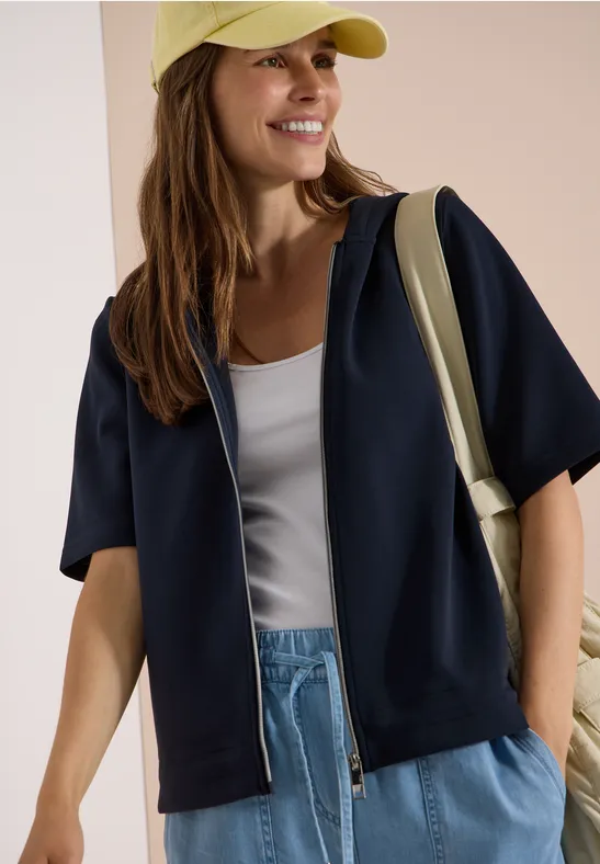 Femme portant une veste à capuche noire, débardeur blanc, jean bleu, casquette jaune et sac à l'épaule.