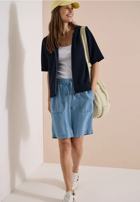 Femme en tenue décontractée: short en jean, haut blanc, veste bleue, baskets, casquette jaune et sac en toile.