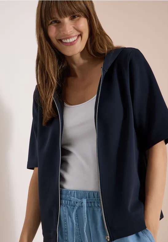 Femme en sweat à capuche bleu à manches courtes, débardeur blanc et jean, visage flouté.