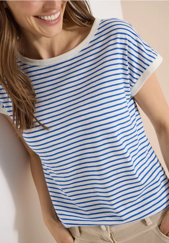 Eine lächelnde Frau lehnt an der Wand, trägt ein blau-weiß gestreiftes T-Shirt und helle Hose.
