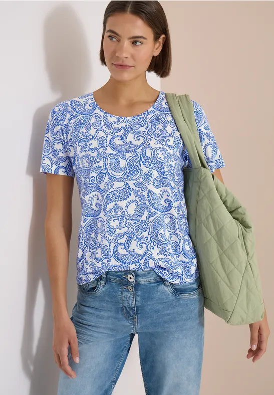 Femme portant un t-shirt imprimé bleu et blanc, un jean et un sac matelassé vert.