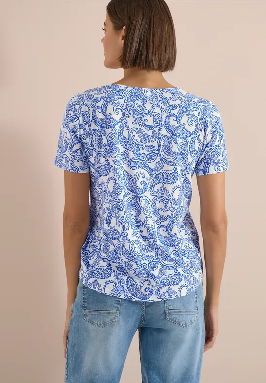 Vue de dos d’une femme portant un t-shirt court bleu et blanc à motifs et un jean bleu clair sur fond beige.