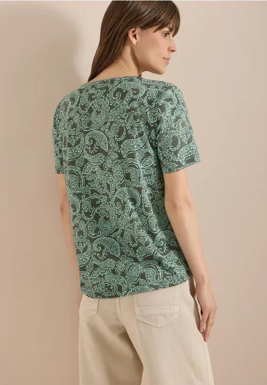 Frau von hinten trägt grünes Paisley-T-Shirt und beige Hose vor hellem, neutralem Hintergrund.