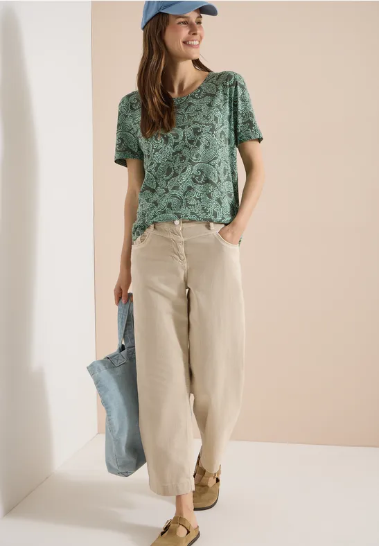 Frau trägt grün gemustertes T-Shirt, beige Hose, blaue Tasche und Cap.