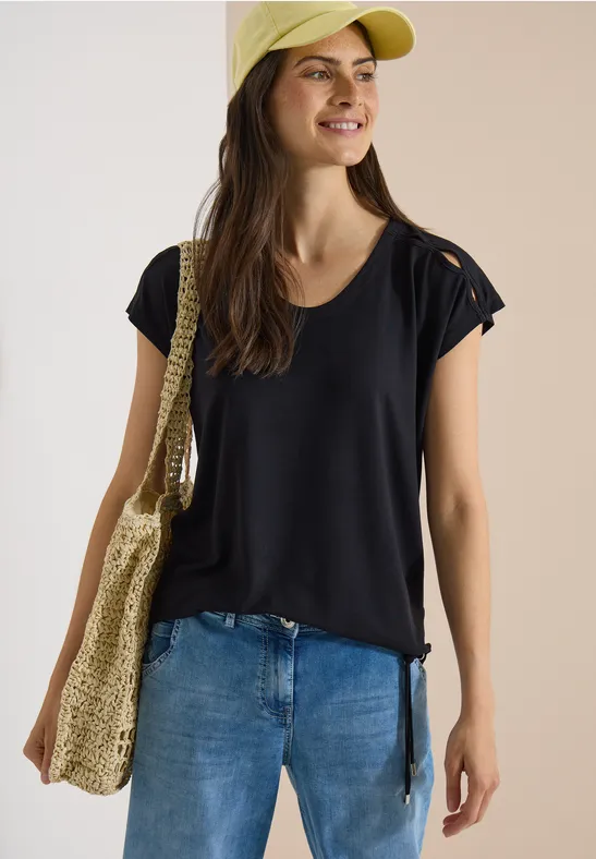 Personne en T-shirt noir, jean, casquette jaune et sac en paille, posant devant un fond neutre.