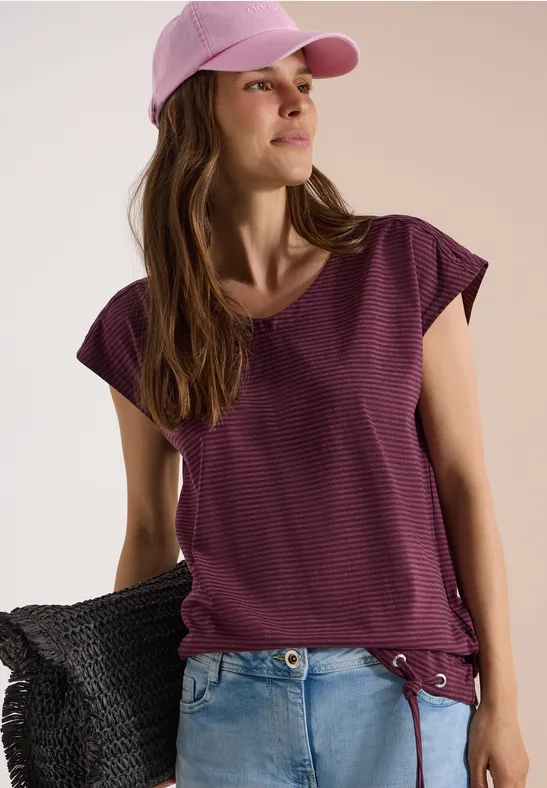 Jeune femme en jean et t-shirt violet à rayures, casquette rose et sac noir, sur fond clair.
