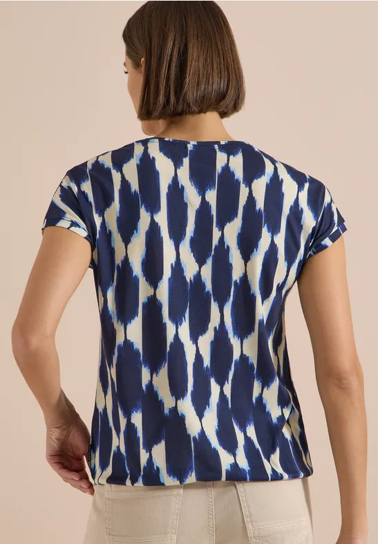 Rückansicht einer Frau in kurzärmeligem T-Shirt mit blau-weißem, abstraktem Musterdruck und heller Hose.