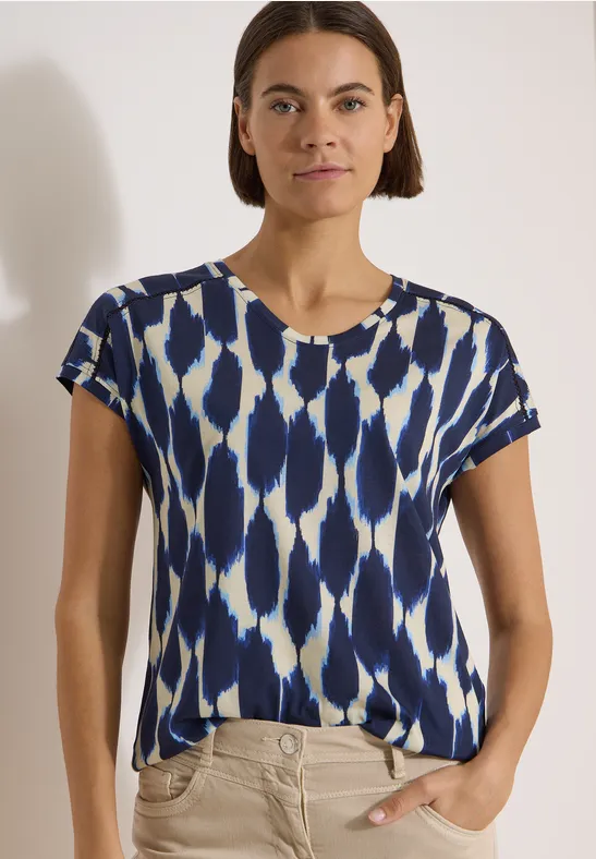 Frau in kurzärmligem Shirt mit blau-weißem abstraktem Muster, eine Hand in Tasche, weißer Hintergrund.