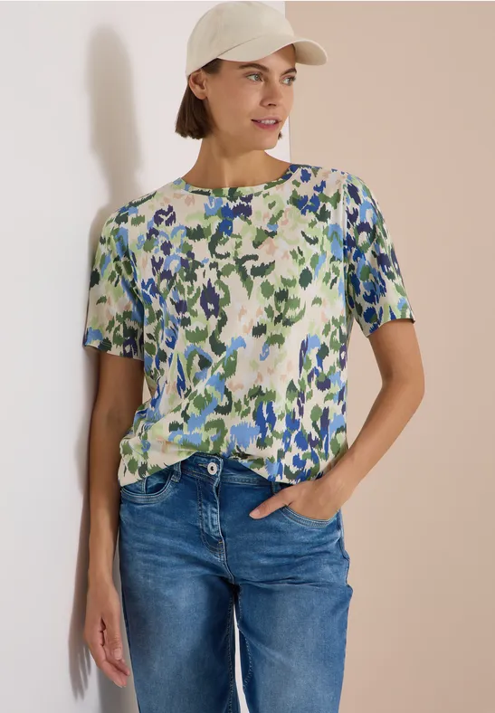 Femme portant un t-shirt fleuri, un jean et une casquette crème sur fond clair.