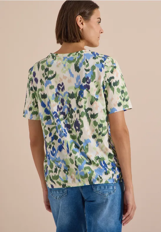 Femme vue de dos, cheveux courts, portant un T-shirt vert et bleu à motifs et un jean bleu.