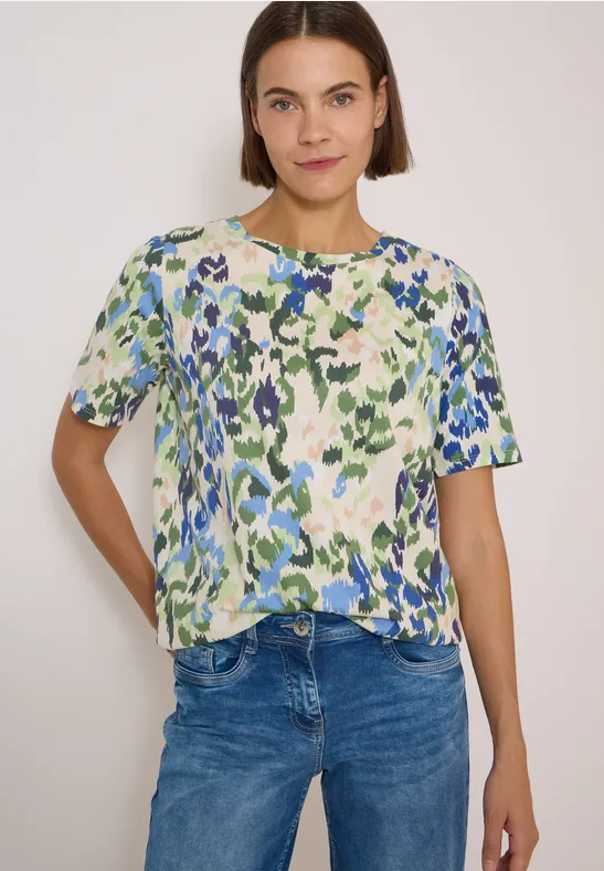 Femme portant un T-shirt à manches courtes coloré à motifs floraux abstraits vert-bleu et jean.