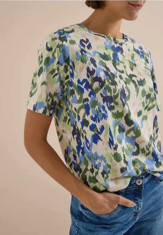 Femme portant un chemisier à manches courtes à motif floral abstrait bleu et vert, jeans bleu.