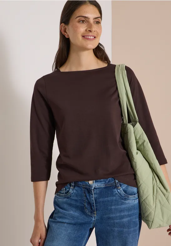 Frau in braunem Dreiviertelarmshirt, Jeans und grüner Stepp-Tasche auf schulter, lächelnd.