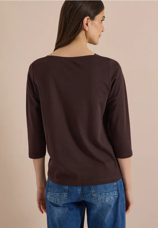 Frau von hinten in braunem Dreiviertelarmshirt und Jeans vor beigem Hintergrund.