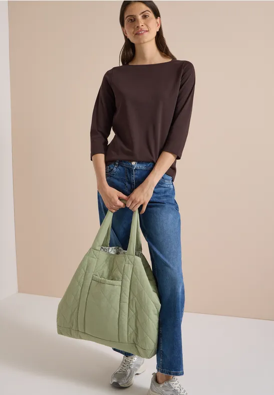 Frau in braunem Shirt und Jeans trägt eine hellgrüne, gesteppte Tasche und weiße Sneaker.