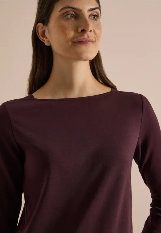 Porträt einer Frau im lila Langarmshirt, die seitlich in die Kamera blickt vor neutralem Hintergrund.
