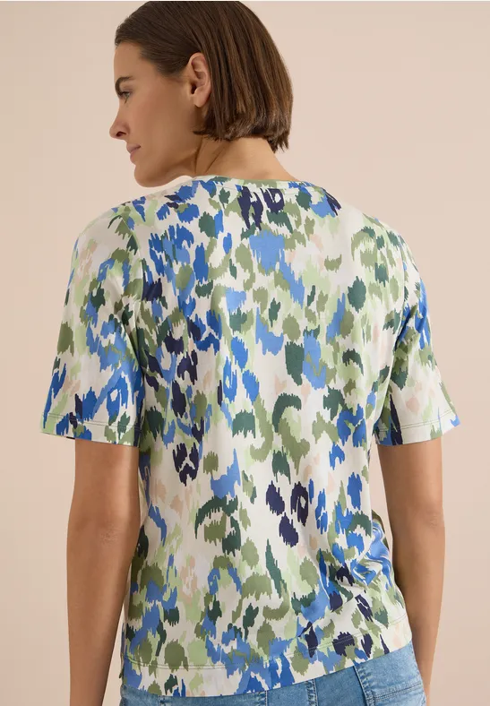 Rückansicht einer Frau mit kurzem braunem Haar und einer kurzärmligen Bluse mit grün-blauem abstraktem Muster.