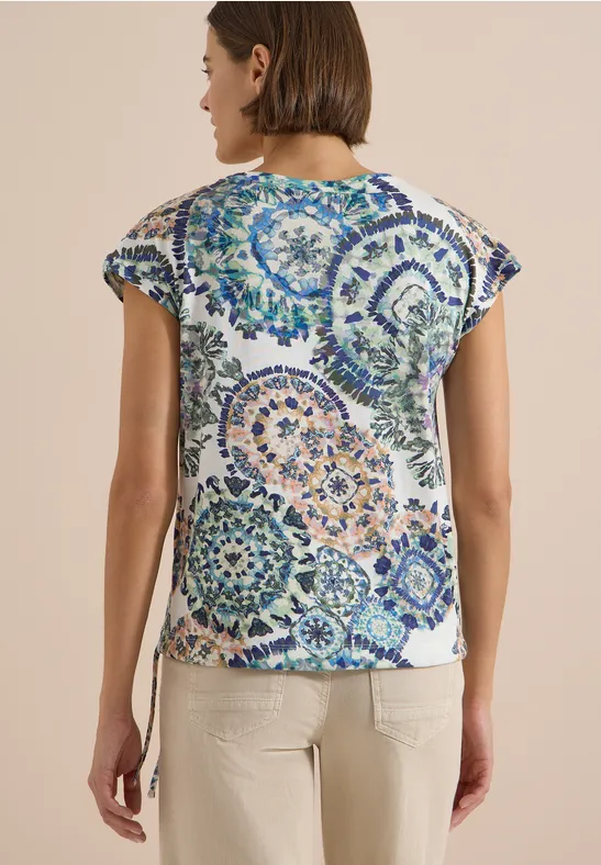 Femme de dos portant un t-shirt blanc à motifs ronds mandala colorés et un pantalon beige.