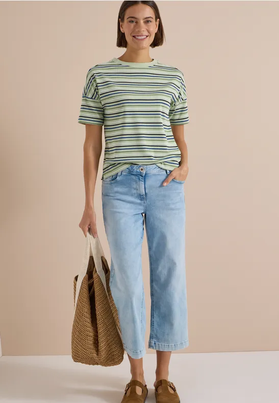 Une femme souriante porte un t-shirt rayé, un jean large et tient un sac en osier.