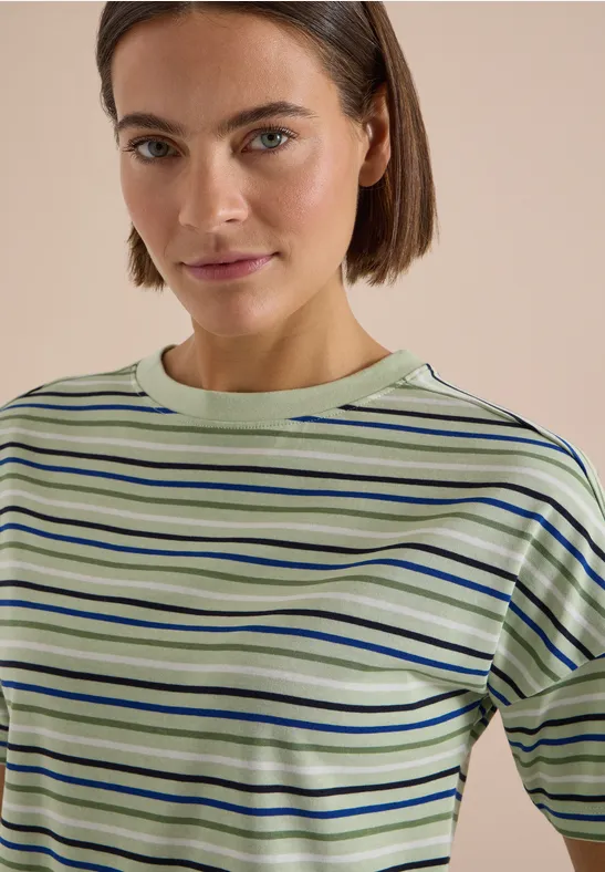 Une femme aux cheveux bruns courts porte un T-shirt vert et bleu à rayures sur fond beige.