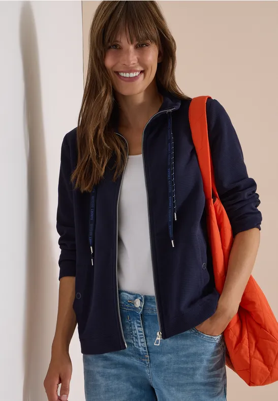 Frau in dunkelblauer Kapuzenjacke, weißem Shirt, Jeans und oranger Tasche lächelnd.