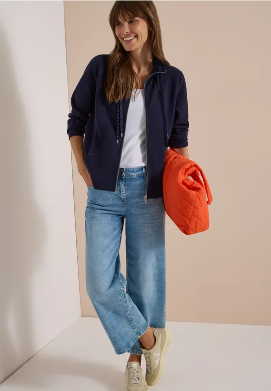Eine lächelnde Frau in Jeans, weißem Shirt, blauer Jacke und Turnschuhen hält einen orangen Pullover.