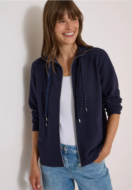 Lächelnde Frau in dunkelblauer Kapuzen-Jacke, weißem Top und Jeans vor hellem Hintergrund.