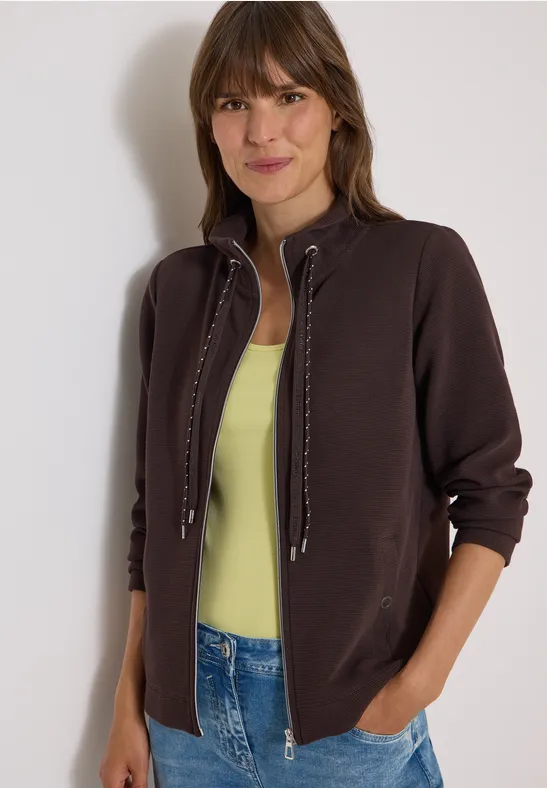 Eine junge Frau lächelt, trägt eine braune Jacke über einem hellgrünen Top und Jeans.