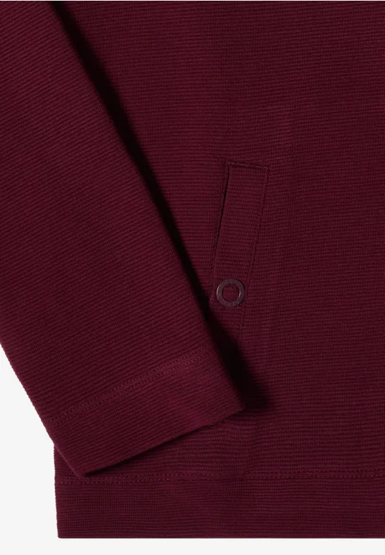 Nahaufnahme eines bordeauxroten Pullovers mit Rippbündchen und aufgesetzter Tasche mit Druckknopf.