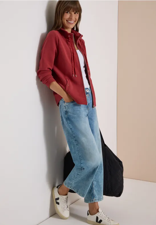 Junge Frau mit roter Jacke, weißem Shirt, weitem Jeans und Sneakern lehnt lächelnd an einer Wand.