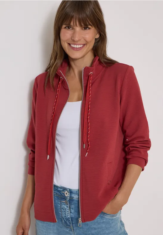 Lächelnde Frau in roter Kapuzenjacke, weißem Shirt und Jeans vor neutralem Hintergrund.