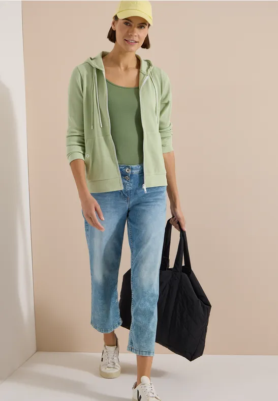 Person trägt grünen Hoodie, grünes Top, weite Jeans und Sneakers, hält schwarze Einkaufstasche.