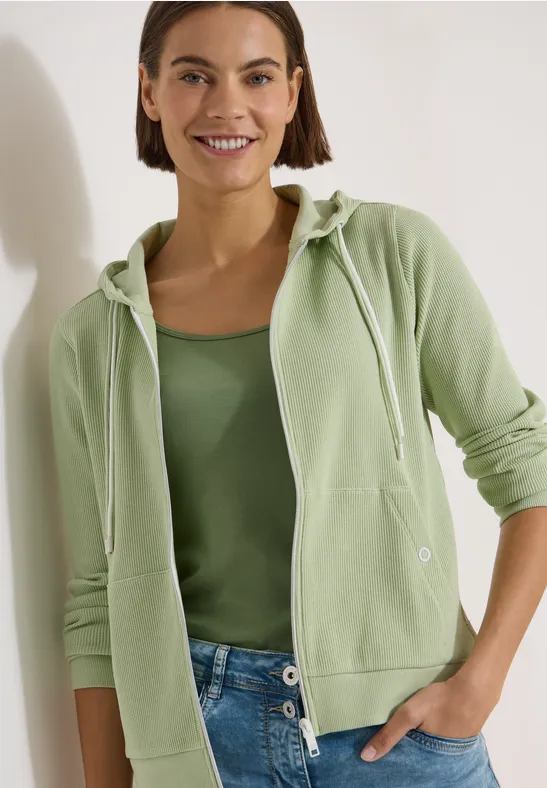 Person in grüner Kapuzenjacke und Tanktop mit blauen Jeans vor hellem Hintergrund.