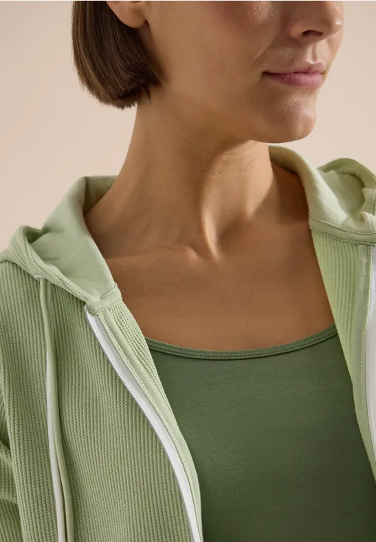 Nahaufnahme einer Person, die ein grünes Tanktop und eine helle Kapuzenjacke trägt.
